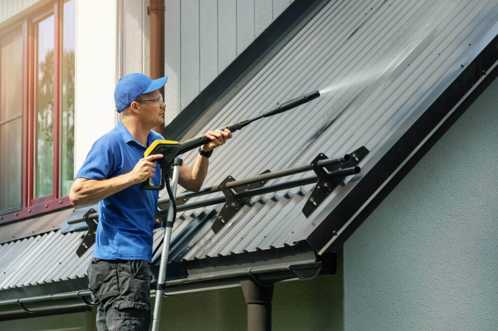 Service worker in a blue shirt and safety glasses pressure washing a corrugated metal roof with solar panel mounts