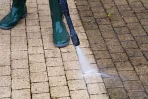 A close-up shot of a person in green rubber boots pressure washing a paving stone walkway, showing a distinct line between the dirty, dark brown stones and the clean, light tan stones.