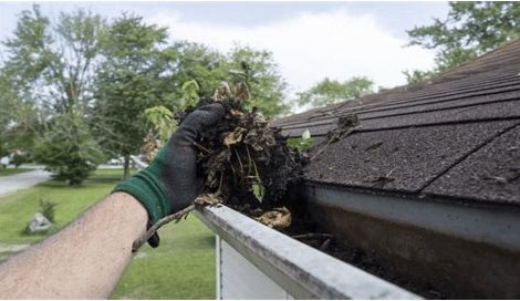 Close-up of a gloved hand pulling a large clump of wet leaves, dirt, and plant debris out of a dirty residential roof gutter.
