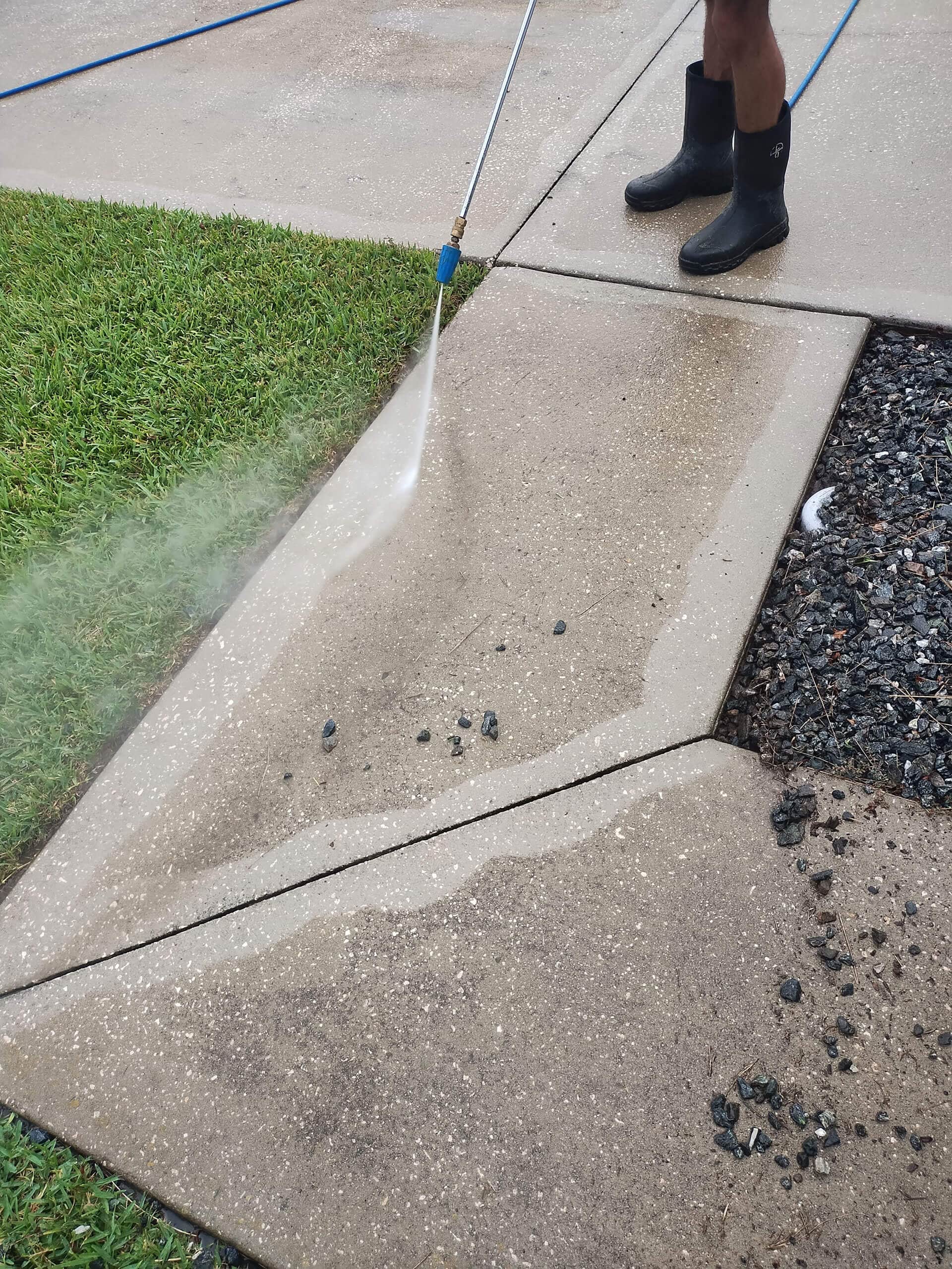 Close-up of a person wearing black rubber boots using a high-pressure washer wand to clean a dirty, exposed aggregate concrete sidewalk or driveway next to a patch of green grass.