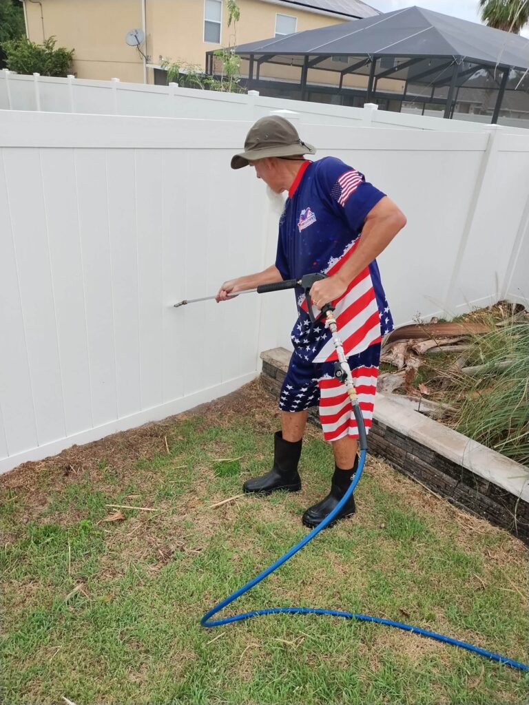 Older man in patriotic clothing pressure washing a vinyl fence, demonstrating professional cleaning techniques for fence maintenance.