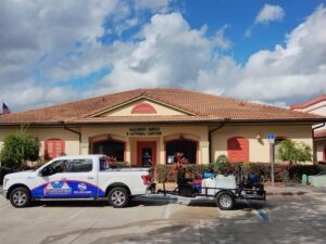White pickup truck branded with a pool and home cleaning and maintenance company logo, towing a trailer with pressure washing and chemical tanks, parked outside a commercial building.