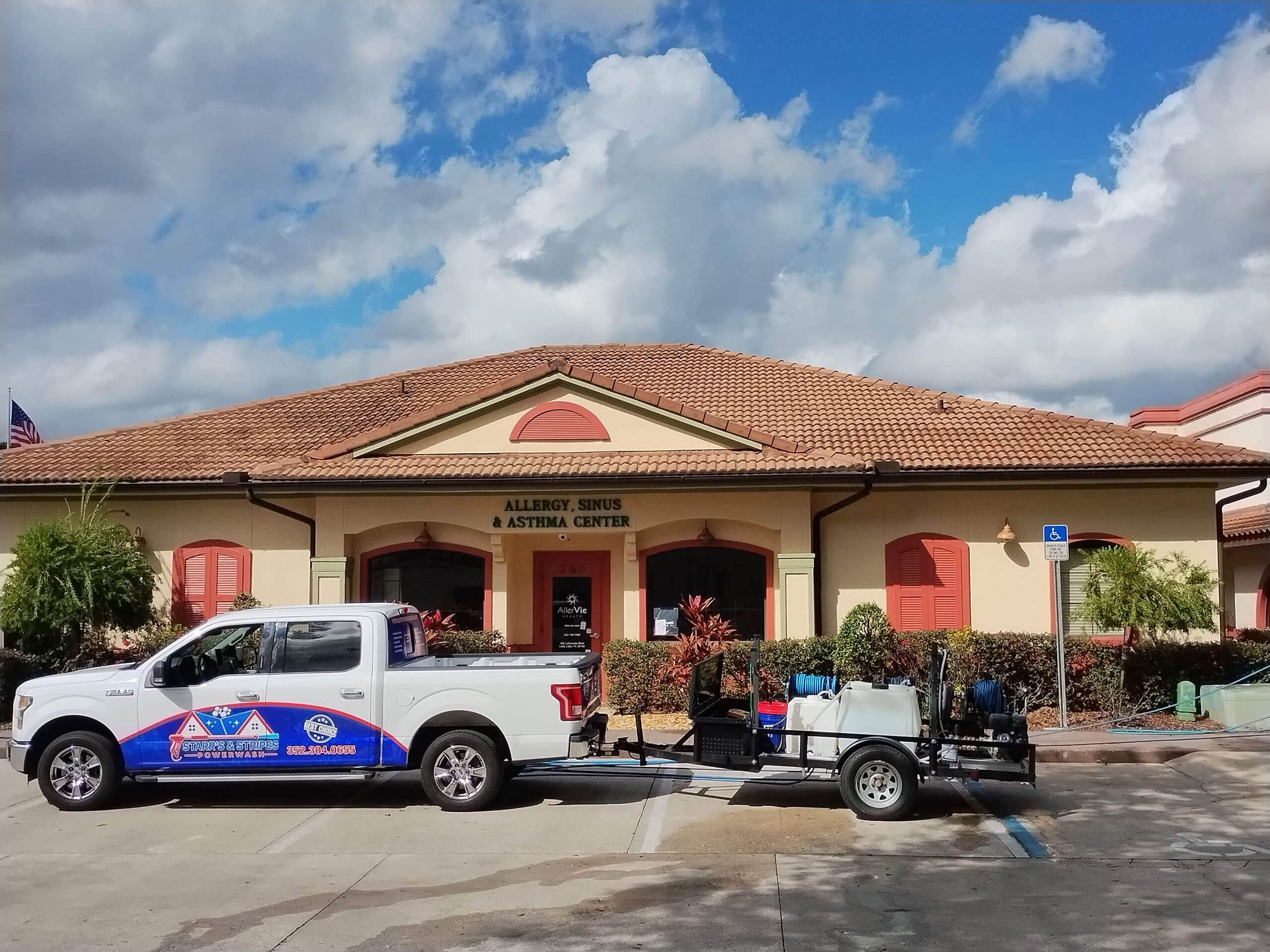 White pickup truck branded with a pool and home cleaning and maintenance company logo, towing a trailer with pressure washing and chemical tanks, parked outside a commercial building.