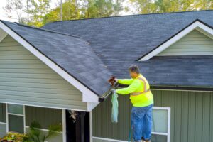 A service worker on a ladder wearing high-visibility safety gear removes leaves and debris from a clogged roof gutter on a residential home.