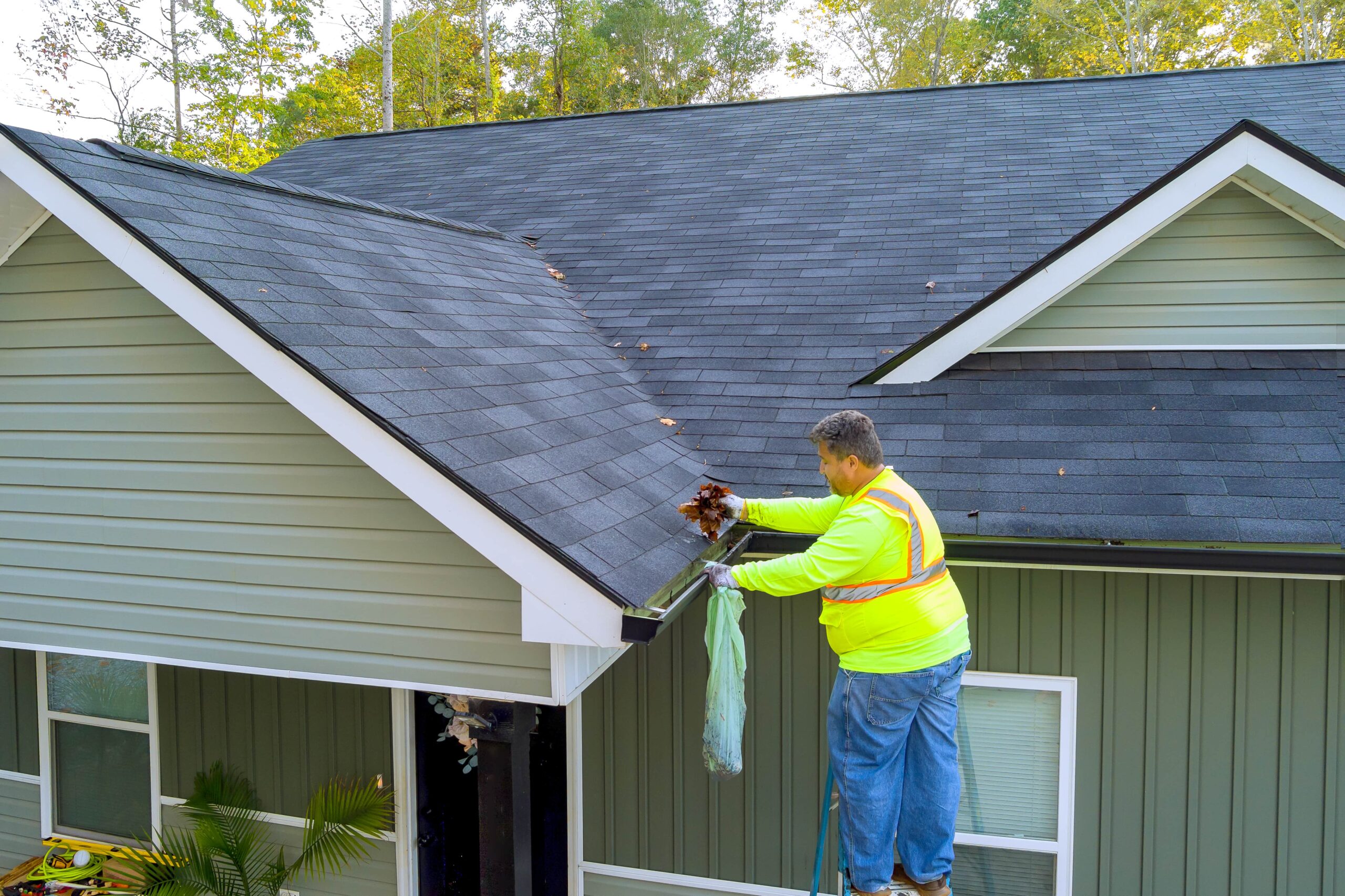 A service worker on a ladder wearing high-visibility safety gear removes leaves and debris from a clogged roof gutter on a residential home.