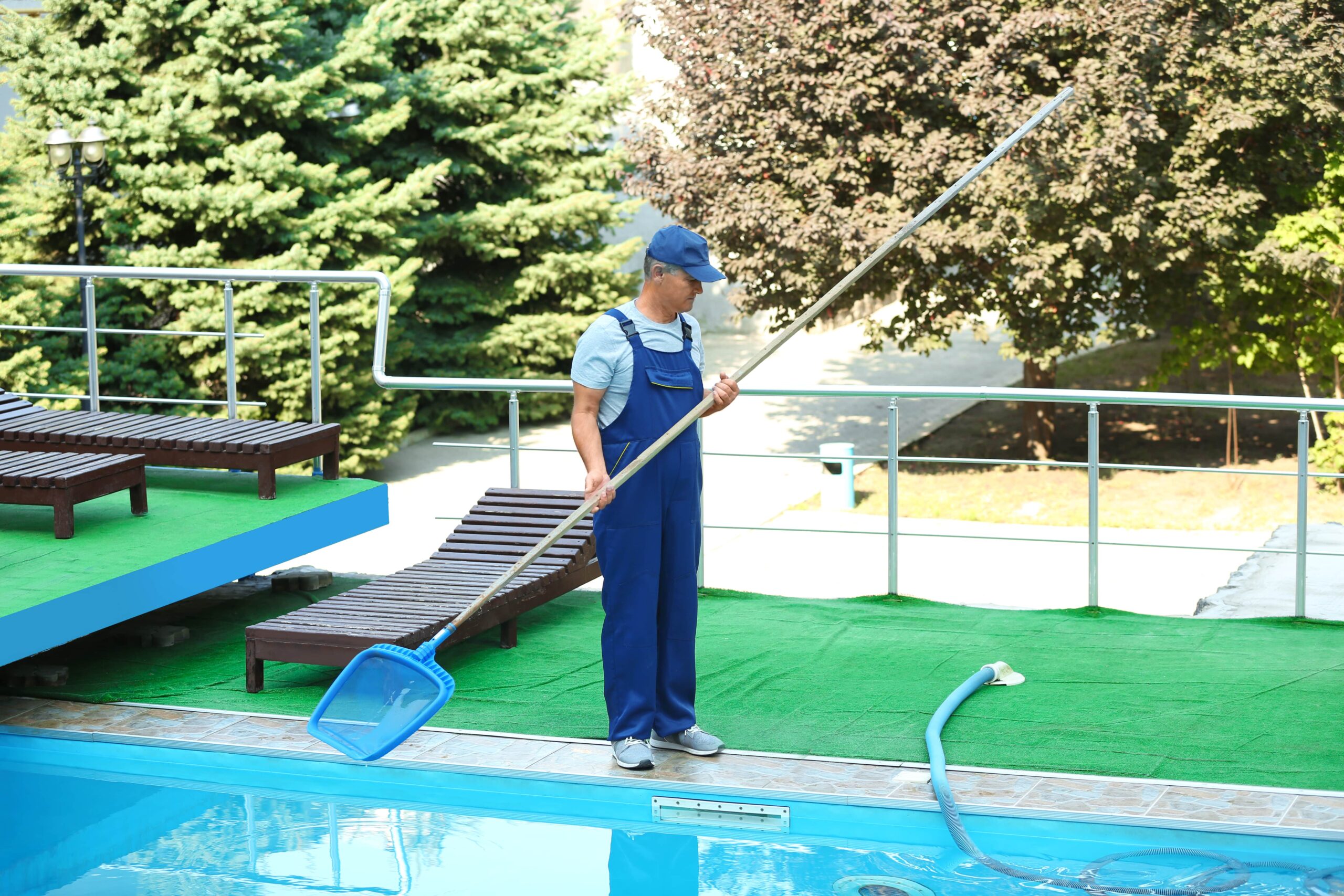 A male pool technician in blue overalls and a cap using a long-handled blue scoop net to clean debris from a clear outdoor swimming pool at a resort or residential property.