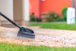 Close-up of a black circular surface cleaner attachment pressure washing a stone paver walkway, removing dirt and debris in a residential backyard.