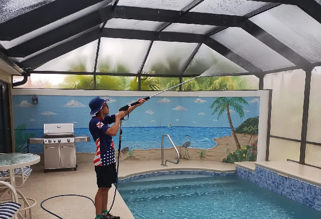 A person pressure washes the ceiling of an enclosed pool area. The scene is bright, with a mural of a beach and palm trees in the background