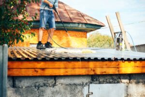 A man in denim shorts stands on a corrugated metal roof, using a high-pressure water sprayer to deep clean a white area rug outdoors.