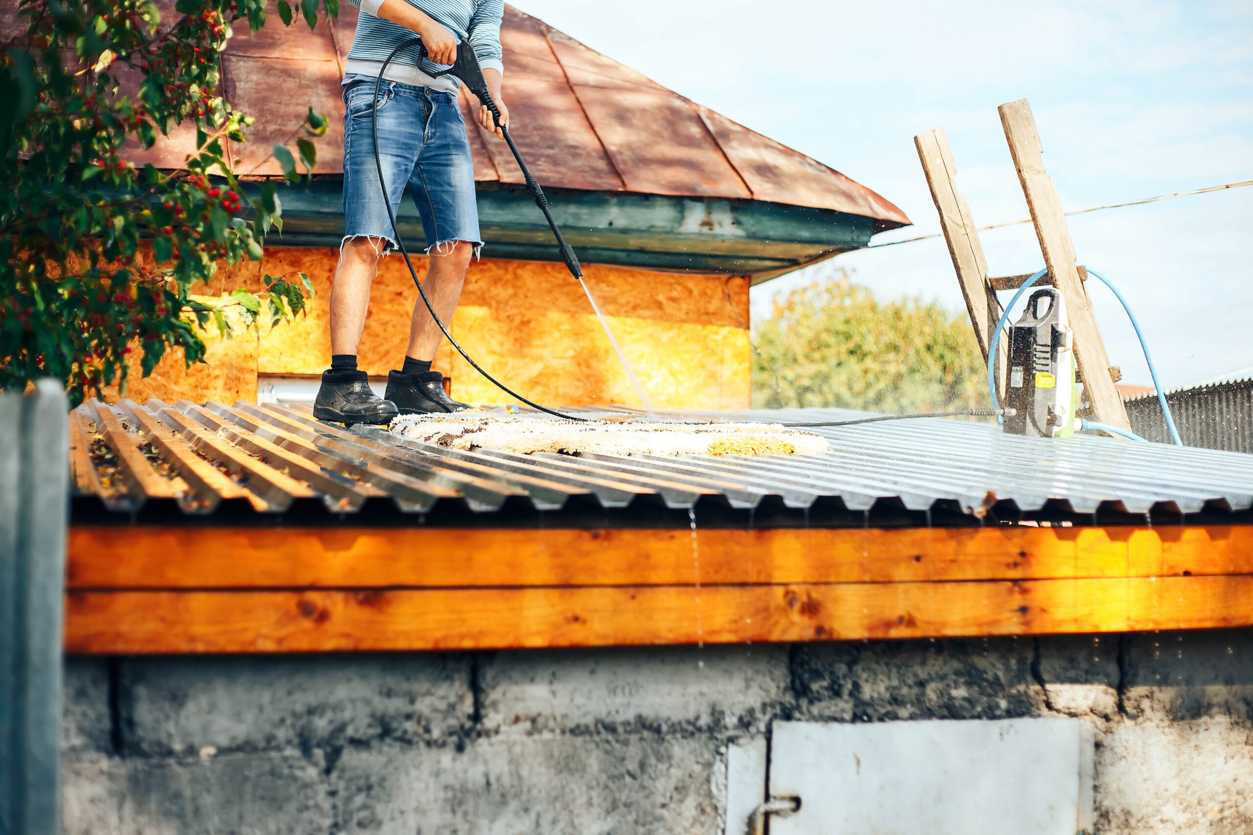 A man in denim shorts stands on a corrugated metal roof, using a high-pressure water sprayer to deep clean a white area rug outdoors.