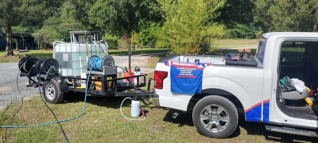 Inverness 9 A white Stars and Stripes Powerwash pickup truck towing a professional pressure washing trailer equipped with a water tank and hose reels, parked on the grass in Ocala, FL. A company shirt draped on the truck lists concrete cleaning, house washing, roof washing, and paver sealing services.