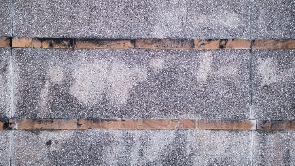 A weathered, textured exposed aggregate concrete wall featuring horizontal brown stone tile accents and dark organic staining, ready for professional concrete pressure washing services in Austin, TX.