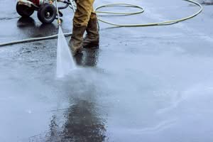 A close-up view of a worker wearing heavy boots and brown pants using a high renting a pressure washer to blast away dirt, oil stains, and debris from a wet asphalt driveway surface in Nashville, TN.