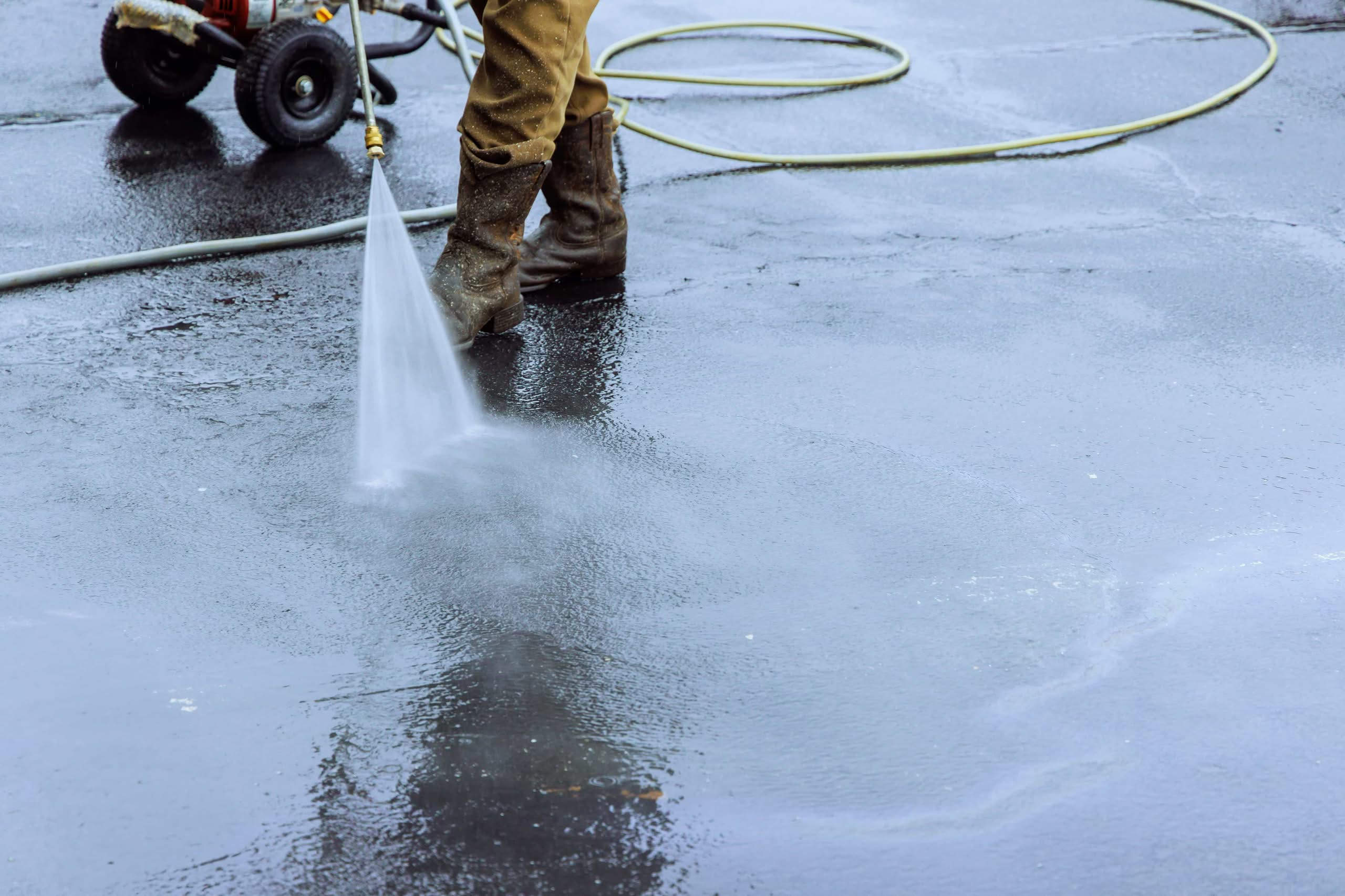 Stop! Renting a Pressure Washer Costs Thousands (2026) 1 A close-up view of a worker wearing heavy boots and brown pants using a high renting a pressure washer to blast away dirt, oil stains, and debris from a wet asphalt driveway surface in Nashville, TN.