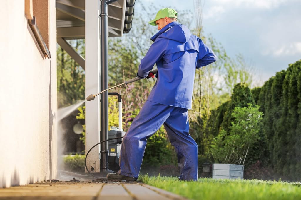 Stop Ruinous Stucco Damage in Ocala: Proven Humidity Solutions 3 A professional technician in blue protective gear and a green cap uses a pressure washer to spray down the stucco damage in Ocala, exterior siding of a house on a bright sunny day in Charlotte, NC.