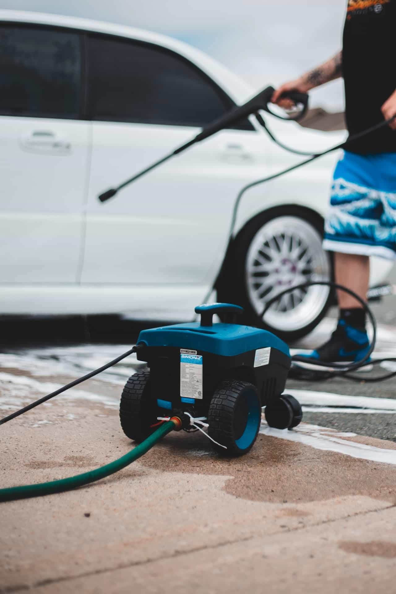 Parking Lot Cleaning 1 A blue Simoniz pressure washer sits on a soapy driveway while a person uses the wand to wash a white sedan, representing professional mobile auto detailing in Los Angeles, CA.