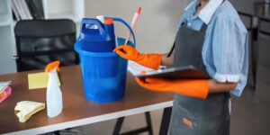 A professional Ocala commercial cleaning in an apron and orange gloves holding a clipboard next to a blue cleaning bucket and supplies on a desk in Atlanta, Georgia.