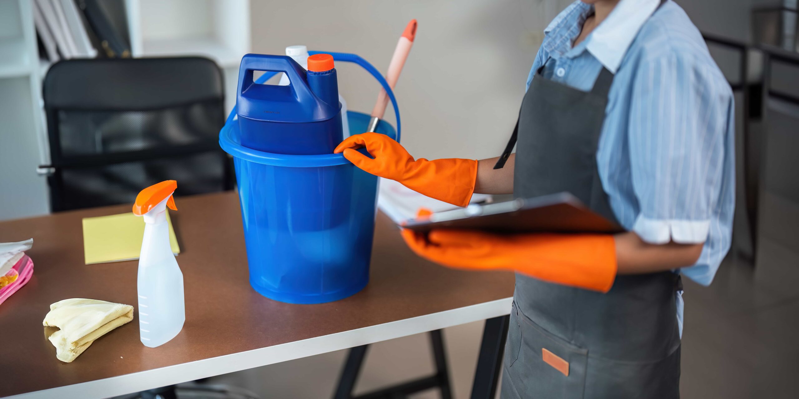 Ocala Commercial Cleaning: Facility Manager's Guide 1 A professional Ocala commercial cleaning in an apron and orange gloves holding a clipboard next to a blue cleaning bucket and supplies on a desk in Atlanta, Georgia.