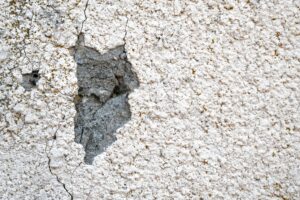 A close-up shot of a textured white stucco exterior wall with a large missing patch revealing the grey concrete base layer underneath, requiring stucco crack repair services in Phoenix, AZ.