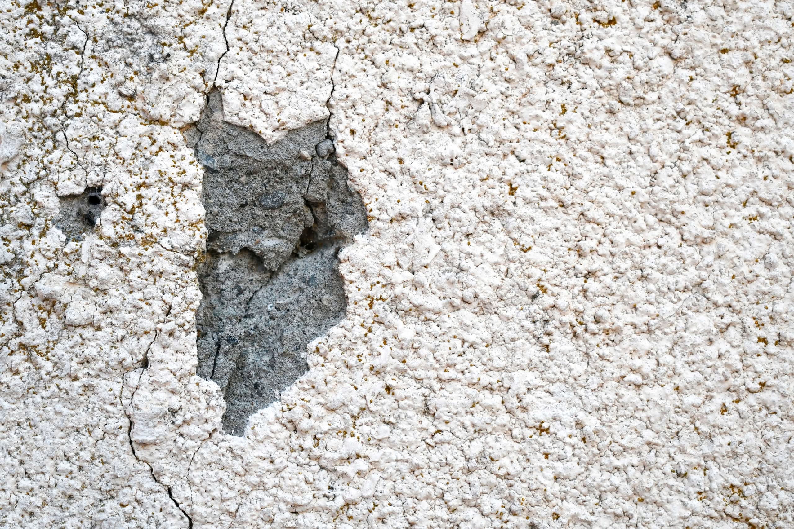 Stop Ruinous Stucco Damage in Ocala: Proven Humidity Solutions 1 A close-up shot of a textured white stucco exterior wall with a large missing patch revealing the grey concrete base layer underneath, requiring stucco crack repair services in Phoenix, AZ.