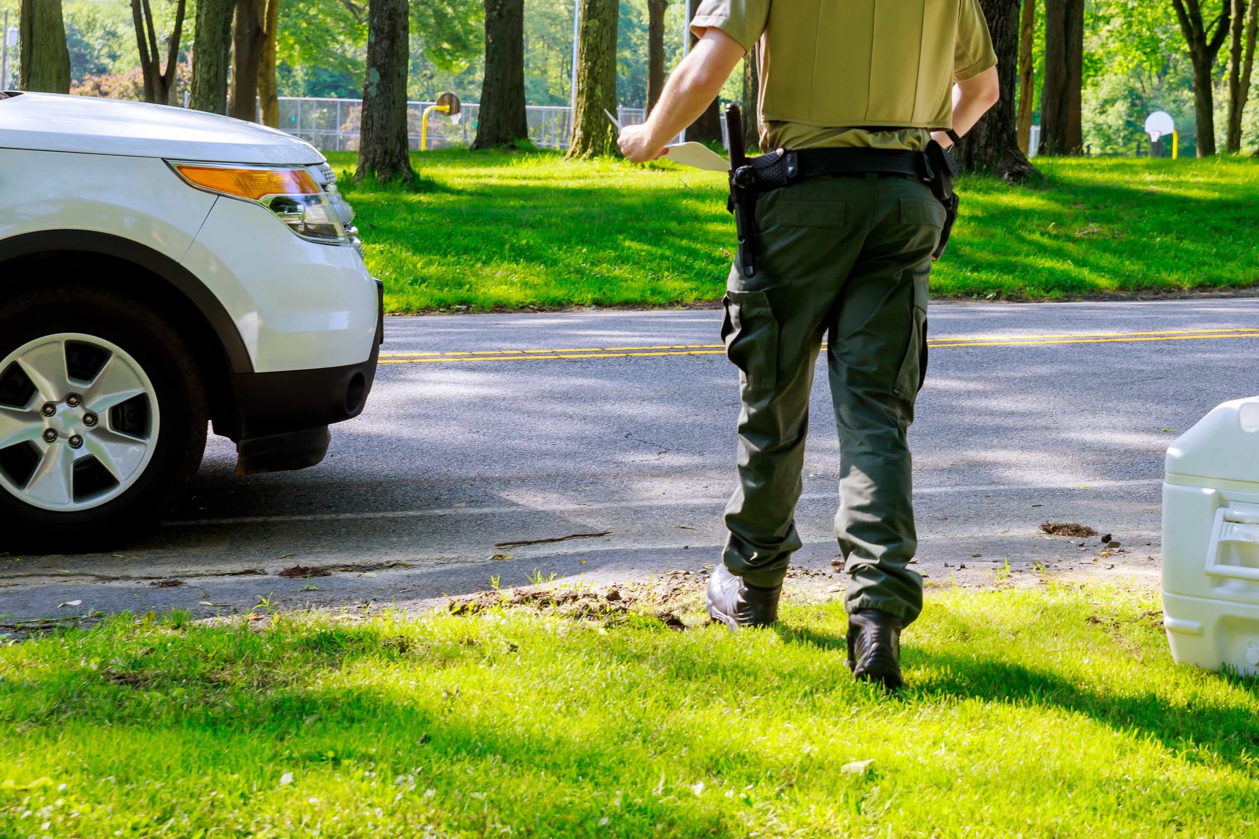 A rear view of a community enforcement officer or security officer in uniform holding paperwork while walking along a driveway toward a parked white vehicle, representing the risk of HOA Fines in The Villages: Avoid Costly Driveway Violations.