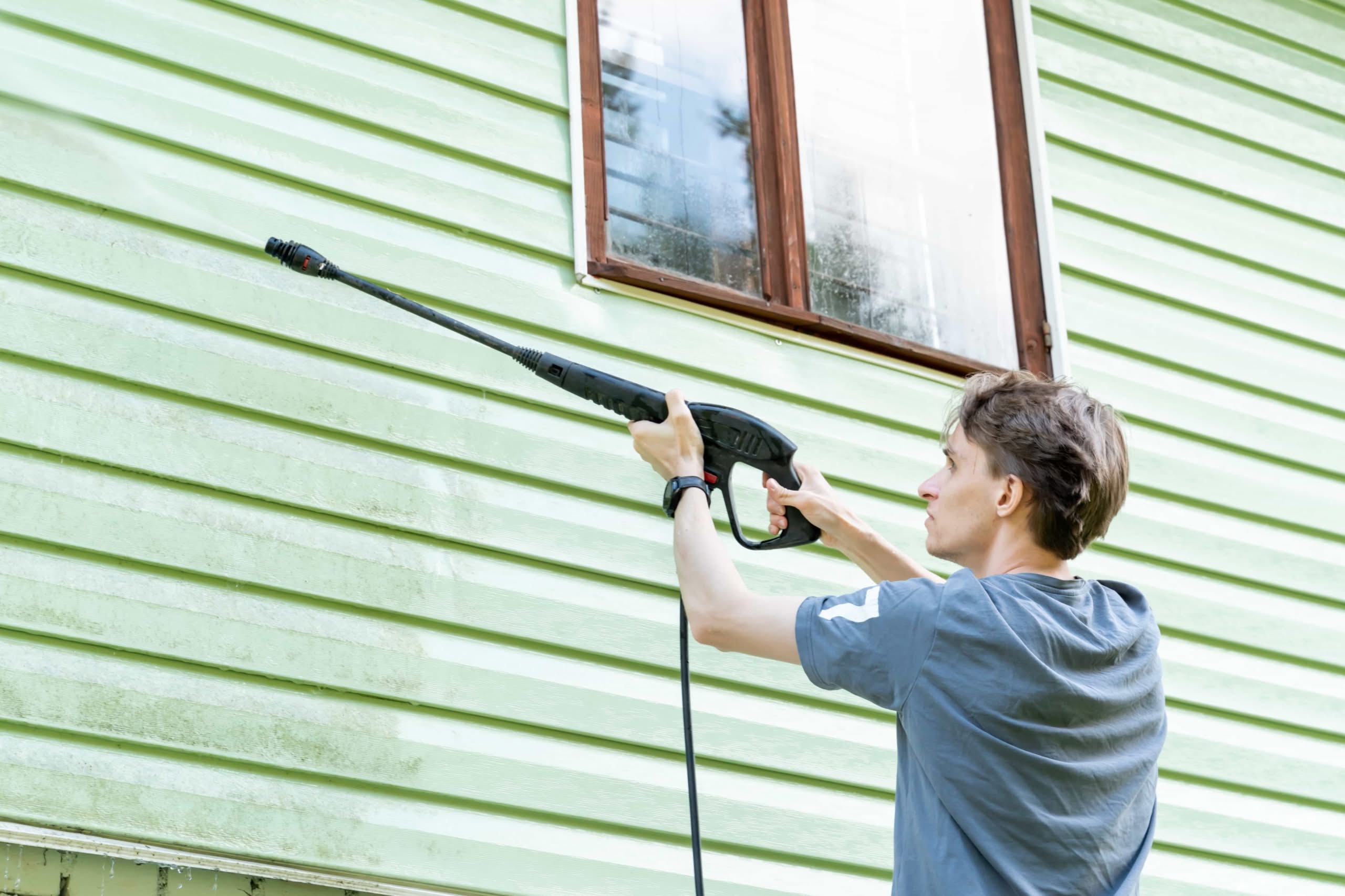 Mobile Home Wash Ocala: Proven Guide to a Spotless Exterior 1 A man standing outdoors using a long pressure washer wand to spray down light green vinyl siding next to a window during a mobile home wash in Mesa, AZ.