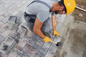 A professional contractor wearing a yellow hard hat and gloves installs stone brick pavers on a driveway base using a rubber mallet, optimized for paver sealing, weeds Between Pavers in Ocala, Florida.