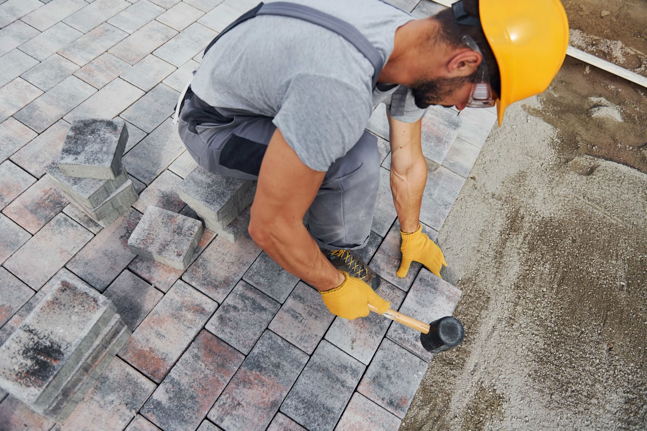 Weeds Between Pavers: Why They Keep Coming Back in Florida 1 A professional contractor wearing a yellow hard hat and gloves installs stone brick pavers on a driveway base using a rubber mallet, optimized for paver sealing in Orlando, FL.