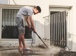 A man in casual clothing using a pressure washer wand to clean a dirty concrete patio surface, illustrating the problem of how rented pressure washers leave streaks on concrete and the fix needed in Houston, TX.
