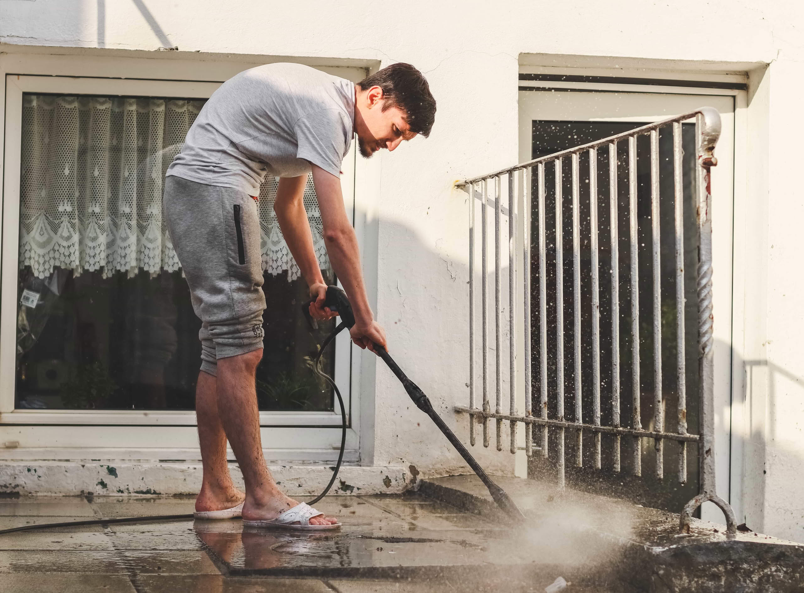 A man in casual clothing using a pressure washer wand to clean a dirty concrete patio surface, illustrating the problem of how rented pressure washers leave streaks on concrete and the fix needed in Houston, TX.